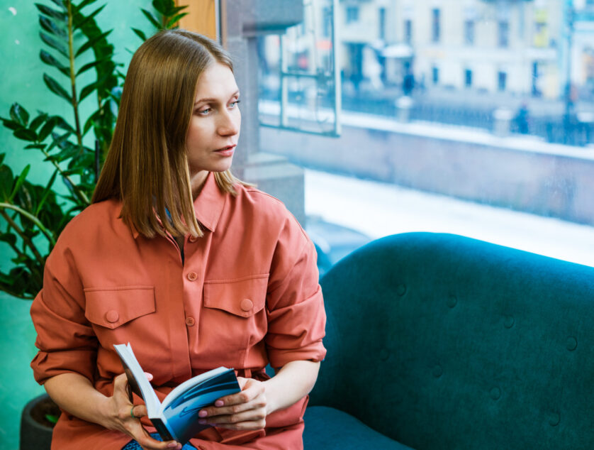 Beautiful caucasian girl spends her free time at home, sitting on couch, reading a book and looking thoughtfully out the window. Caucasian woman lifestyle, leisure at home